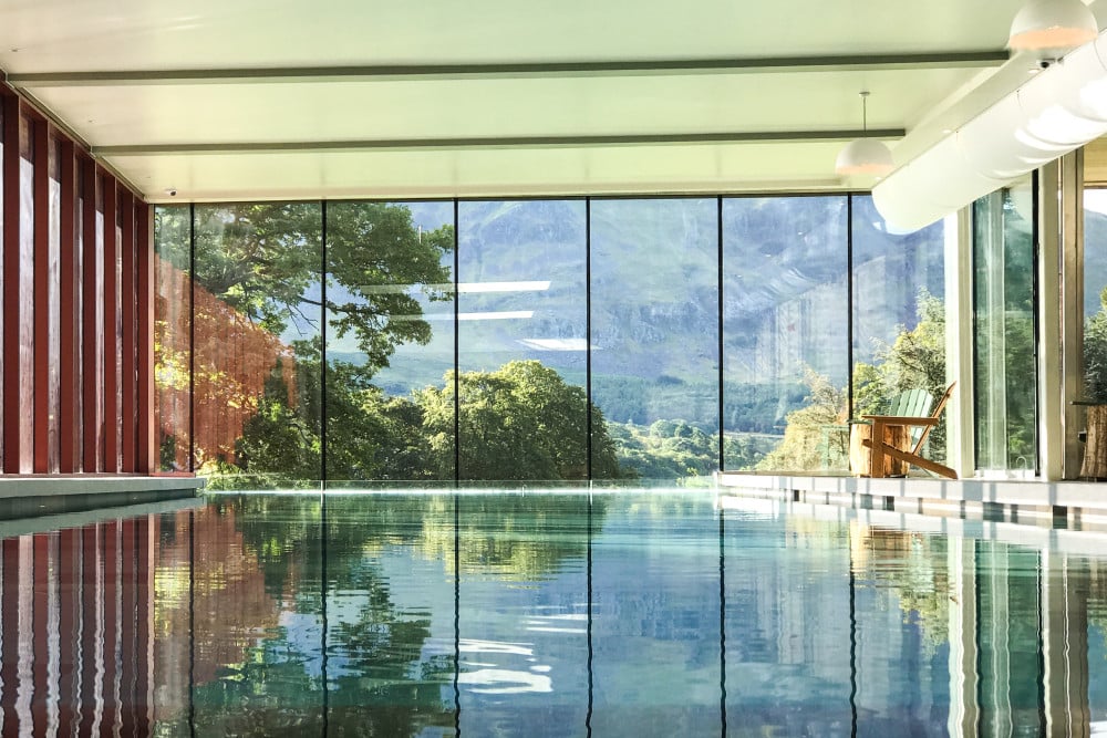 Stunning infinity-edge pool at Another Place The Lake on the shores of Ullswater, featuring floor-to-ceiling glass walls with breathtaking Lake District mountain and valley views, modern minimalist design with exposed beams, and seamless indoor-outdoor spa experience in a luxury countryside retreat