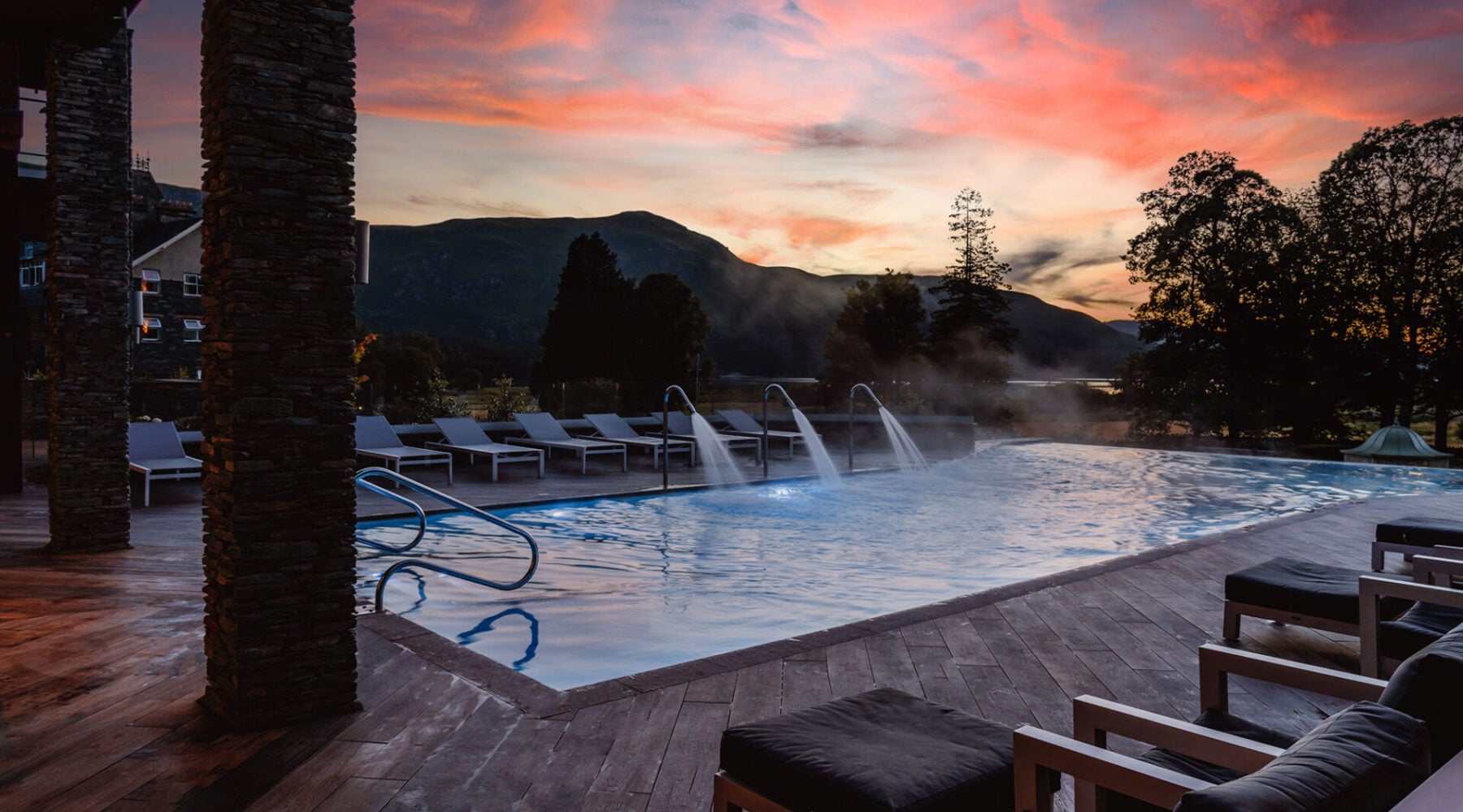 Luxurious 16-metre outdoor hydrotherapy pool at The Falls Spa at Lodore Falls Hotel in Borrowdale, featuring heated water with neck jets and underwater bubble loungers, surrounded by white sun loungers on slate decking with dramatic Lake District mountain views at sunset, spa access included with suite bookings